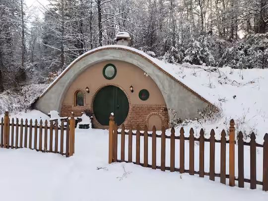A charming hobbit house Airbnb in Virginia during winter, featuring a green round door and a rustic wooden fence.