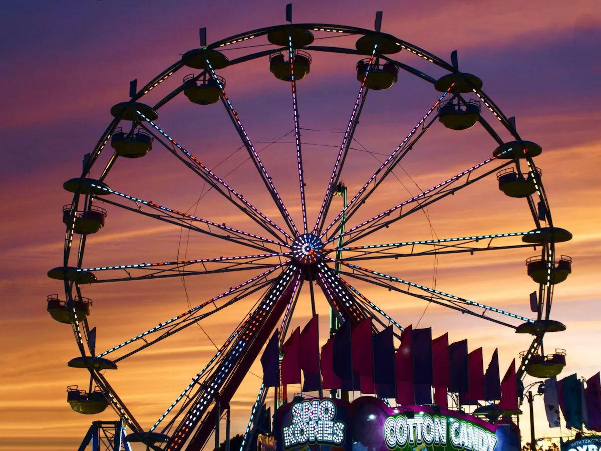 A colorful Ferris wheel at the Loudoun County Fair is silhouetted against a vibrant orange and purple sunset sky, with a cotton candy sign visible at the bottom.