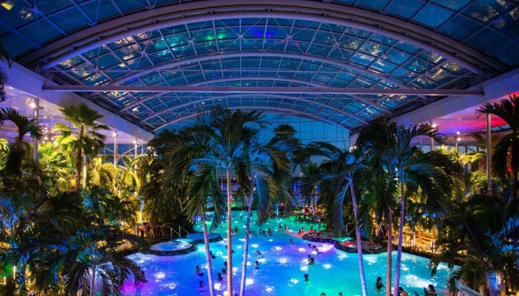 Vibrant night scene inside an expansive Therme thermal resort, featuring a large pool with swimmers, illuminated tropical palm trees, and a glass-domed roof.